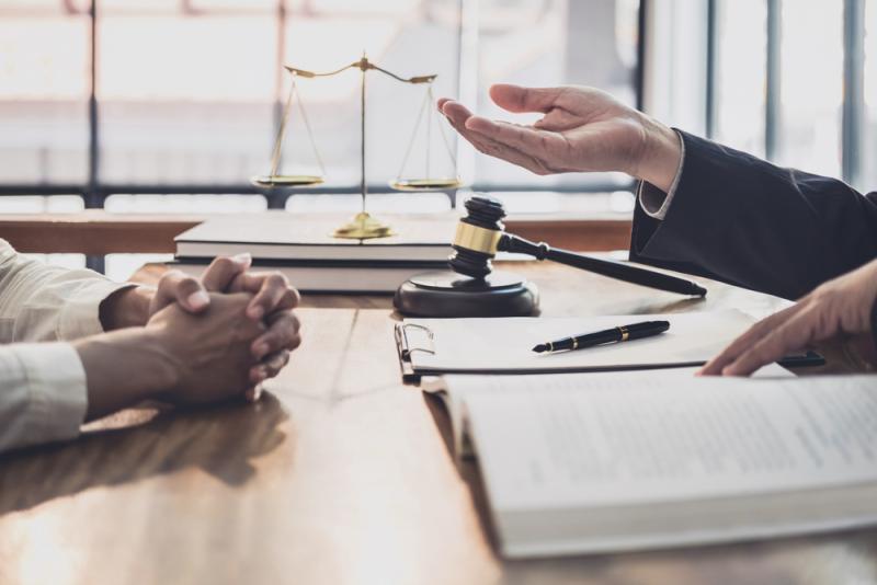 A closeup of two pairs of hands - one pair of hands is clasped, as though listening, the other pair is gesturing to a book of law with a gavel next to it.