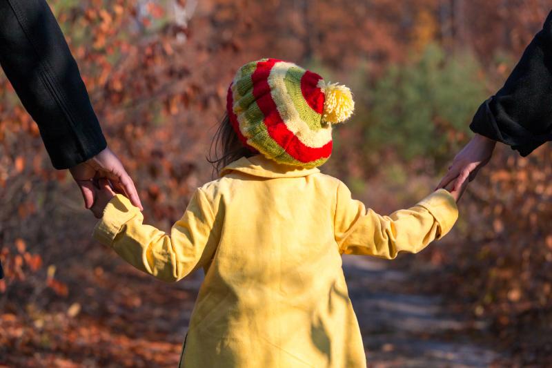 A Caucasian child in a yellow coat holding its parents' hands