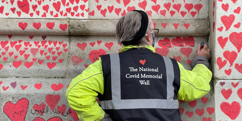 A National Covid Memorial Wall volunteer stands facing the wall as she writes a message. She wears a 'National Covid Memorial Wall' vest.