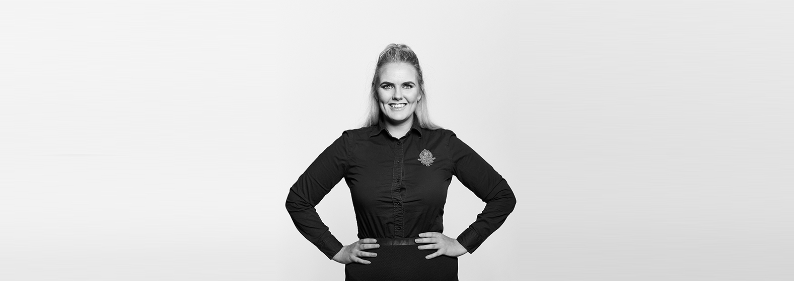 A grayscale photo of Michaela O'Brien, Legal Executive. She is wearing a dark long-sleeved top and has her hands on her hips and is smiling.