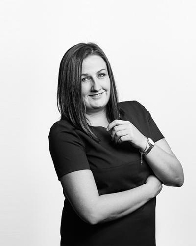A grayscale photo of Danielle Carter, Associate Solicitor. She wears a dark short-sleeved blouse and has her arms crossed slightly, she is smiling.