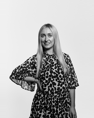 A greyscale photo of Grace McNamara. She has long blonde hair, is wearing a black blazer and a white blouse. She is smiling