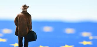 A photo of a small model figure of a person in a brown coat and brown hat carrying a suitcase facing away from the camera. The floor the figure is standing on is the European Union flag and it looks like the figure is looking at the flag.