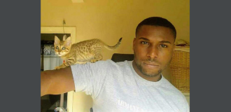 Gavin Brown, a Black man with a fade wearing a white tshirt, is holding out his right arm to the side. A small stripy kitten is balancing on his outstretched arm.