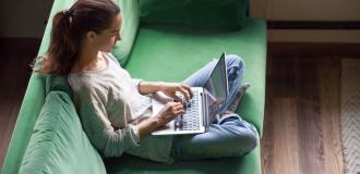 A Caucasian woman sitting on a green sofa with her laptop on her lap.