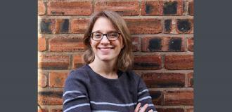 Charlotte Mills stands against a brick wall. Her arms are folded and she is smiling assuredly.