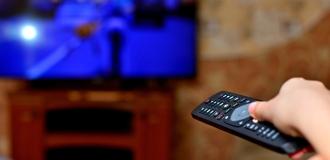 A close-up of a caucasian hand holding a TV remote, pointing it at a television in the background