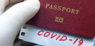 A hand wearing a medical glove holds a burgundy passport. A slip of paper is peeking out of the passport, it reads 'Covid-19' in red writing.