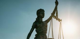 A bronze statue of Justice holding balancing scales up in one hand, the other hand extended behind her. She is in front of a blue sky and is backlit by the sun.