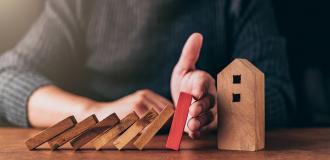 A series of wooden blocks that have fallen into each other. A white person puts a hand between the last block and a wooden model of a house. 