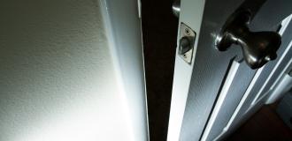 A down-angled close up of a door that is slightly ajar. The hallway beyond the door cannot be looked into, the photo is very shadowy and looks menacing.