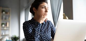 A young woman with black hair wearing a dark blue shirt sits in front of a laptop. She looks off into the distance pensively. 