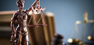 A small bronze statue of Justice holding the scales and the sword is in the foreground. There are books and a set of scales in the blurry background.