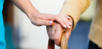 A carer supports the an elderly person's hand. They are holding a cane.