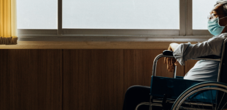 An elderly person in a wheelchair sits gazing out of a window with a medical mask on their face