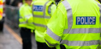 Three white police officers facing away from the camera, slightly out of focus. They are wearing bright yellow high-vis coats with 'Police' written on the back.