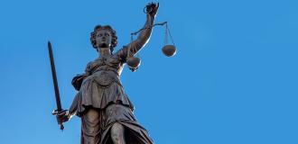 A bronze statue of Lady Justice against a clear blue sky. She is holding a pair of scales in one hand and a sword in the other and is blindfolded.