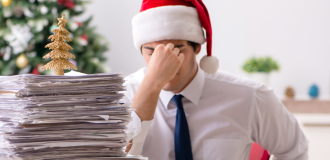 a man wearing a santa hat, holding his head in his hands with a stack of papers on a desk in front of him 