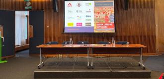 The 'Learn Lessons, Save Lives' report press conference. Two tables stood next to each other in a row with five microphones on them, and five chairs behind the tables. In the background is a projector screen showing the front over of the report and logos of organisations involved, including Broudie Jackson Canter and eight others.