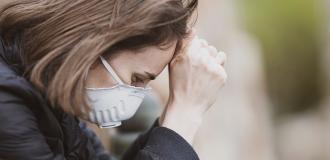 A white women in distress stands leaning on a wall with her head in her hands. She wears a white dust/medical mask.