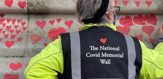 An elderly white woman wearing a 'Covid Memorial Wall' high vis jacket draws a red heart on the Covid Memorial Wall