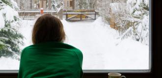 A person with brown hair and an emerald green jumper can be seen from behind, looking out of a large window onto a snowy back garden. There is a cup of tea next to them. 