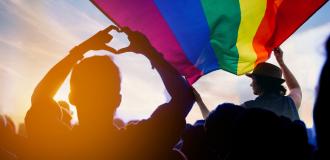A silhouette of a person making a heart with their hands under a large rainbow flag that's held by someone in the foreground.