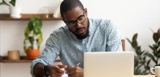 A Black man in a blue shirt sits in front of a laptop and looks at it seriously while making notes on a piece of paper.