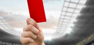 A red card is held up to a cloudy sky on the grounds of a football stadium.