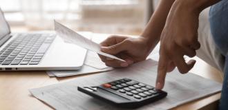 A stack of papers with a calculator on it are on a coffee table. A hand is holding a bit of paper and another hand is pressing a button on the calculator