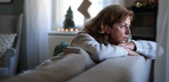 A white woman in her fifties sits on a sofa and leans over the back of it, looking sadly through the window. There are a Christmas decorations on the cabinet behind her. 