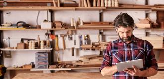 A white man stands in a workshop with carpentry equipment behind him. He's wearing a red and blue plaid shirt and is using an iPad.