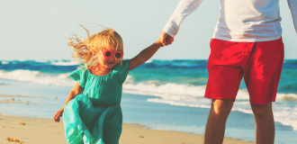 A father holds his daughters hand as they stroll towards the camera along the beach. 