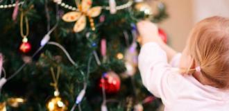 A female white child with blonde hair in pigtails reaches towards a Christmas tree to place a decoration on the branch.