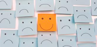 An orange post-it note with a happy face on it amongst many blue post-it notes with sad faces drawn on them