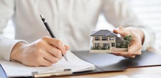 Man with a small model house in his right hand whilst signing a contract with his left hand