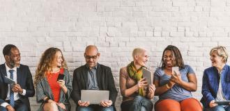 a diverse group of employees sat together on a bench looking at phones, laptops, books