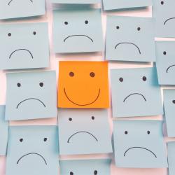 An orange post-it note with a happy face on it amongst many blue post-it notes with sad faces drawn on them