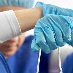 A doctor sits on the floor slumped against the wall. Their arms rest on their knees while their head bows. They're wearing scrubs, blue latex gloves and are holding a mask. 