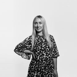 A greyscale photo of Grace McNamara. She has long blonde hair, is wearing a black blazer and a white blouse. She is smiling