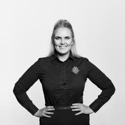 A grayscale photo of Michaela O'Brien, Legal Executive. She is wearing a dark long-sleeved top and has her hands on her hips and is smiling.